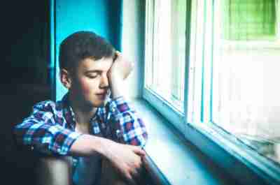 image of young boy feeling stressed with hand against head sitting next to window