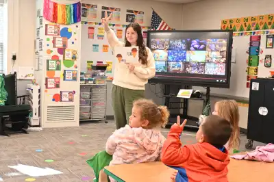 female volunteer in a classroom setting holding flashcard with two ice cream cones, children attentively looking with one student hold their hand up